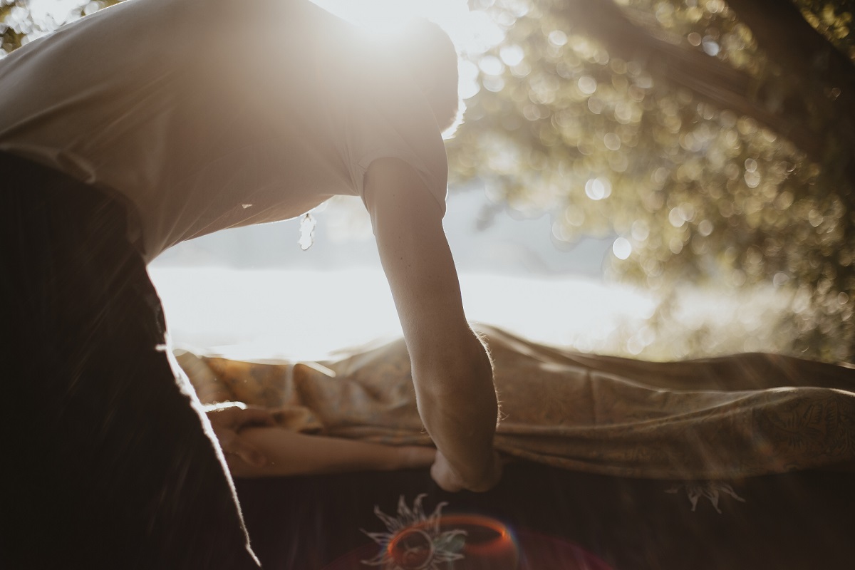 Za masażysty widać delikatnie przebijające się słońce. Pochyla się nad kobietą i wykonuje ruch według masażu lomi lomi nui.